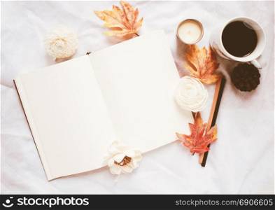 Autumn lifestyle concept, blank notebook and coffee with autumn leaves ornaments on white bed sheet background