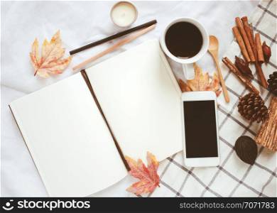 Autumn lifestyle concept, blank notebook, smartphone and coffee with autumn leaves ornaments on white bed sheet background