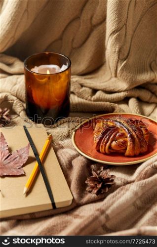 autumn, season and leisure concept - notebook or diary with pencil, cinnamon bun and candle on warm blankets at home. diary, pencil, cinnamon bun and candle in autumn