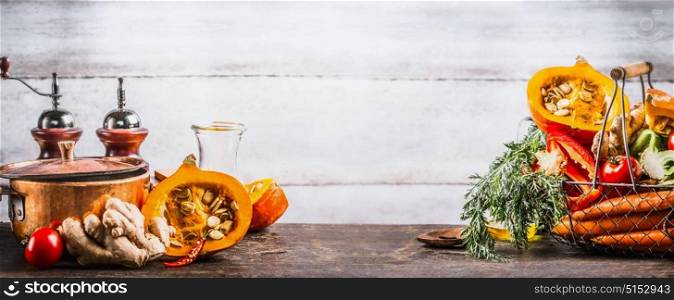 Autumn seasonal cooking concept. Various autumn seasonal organic vegetables: pumpkin,carrot,paprika,tomatoes,ginger in basket on wooden kitchen desk tables with pot at wall background, front view.