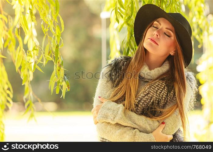 Autumnal female fashion. Pretty young woman wearing stylish sweater waistcoat and hat warm herself outdoor. Beauty fashionable woman standing in park around leaves of willow tree.. Attractive girl warm herself