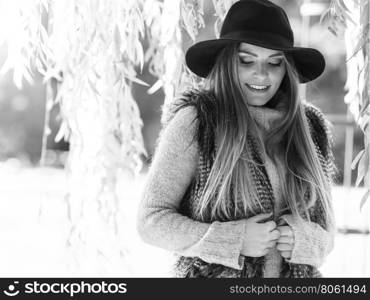 Autumnal female fashion. Pretty young woman wearing stylish sweater waistcoat and hat warm herself outdoor. Beauty fashionable woman standing in park around leaves of willow tree.