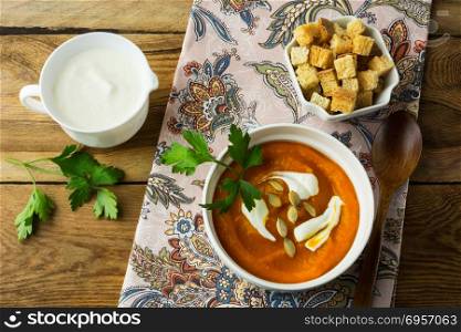 Autumnal spicy pumpkin squash cream soup with cream and pumpkin seeds in the white bowl, cream in a creamer and croutons in a serving dish on the napkin on rustic wooden background, top view. Autumnal spicy pumpkin cream soup