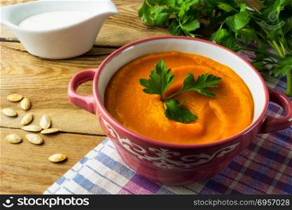 Autumnal spicy roasted pumpkin squash cream soup with parsley in the dark purple-pink soup bowl on the rustic wooden background. Pumpkin soup on the rustic wooden background