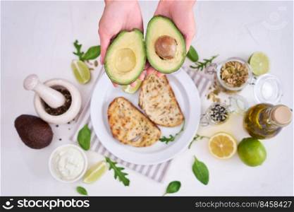 Avocado and cream cheese toasts preparation - Woman holding halved avocado over table with ingredients.. Avocado and cream cheese toasts preparation - Woman holding halved avocado over table with ingredients