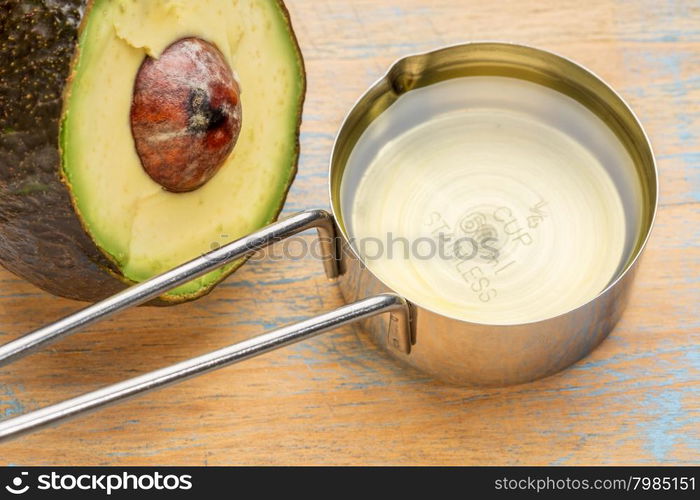 avocado oil in a metal measuring cup against painted wood with a half of avacado fruit