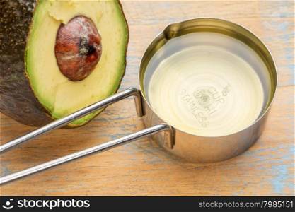 avocado oil in a metal measuring cup against painted wood with a half of avacado fruit