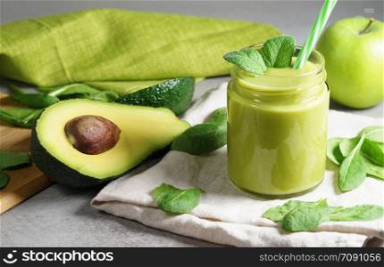 Avocado spinach and green apple smoothie healthy and refreshing tropical drink front view of a glass jar with mint leaves and sliced avocado fruit, apple and spinach leaves on grey kitchen board.