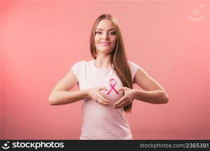 Awareness of breast cancer.Young woman making heart shape by hands on pink ribbon symbol sign. Help and charity. Light red background. Breast cancer. Woman making heart shape on pink ribbon