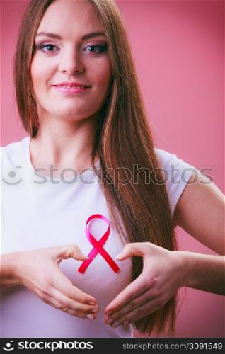Awareness of breast cancer.Young woman making heart shape by hands on pink ribbon symbol sign. Help and charity. Light red background. Breast cancer. Woman making heart shape on pink ribbon