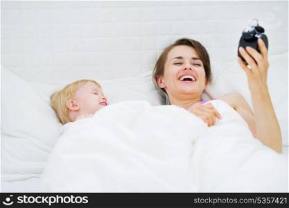 Baby and mother laying on bed and looking on alarm clock