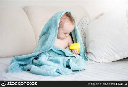Baby boy covered in blue towel playing with yellow rubber duck on sofa after bathing