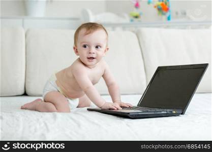 Baby boy in diapers crawling on bed and using laptop com