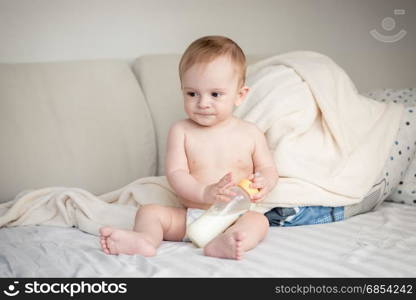 Baby boy in diapers sitting on bed and holding his bottle with milk