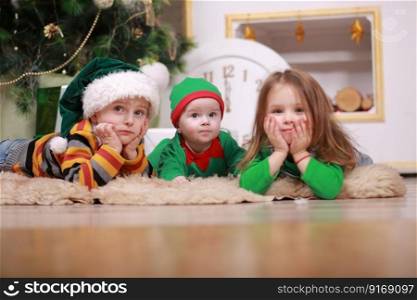 baby boy in red green elf costume with his older brother and sister in santa hats sitting under Christmas tree with gift boxes.. baby boy in red green elf costume with his older brother and sister in santa hats sitting under Christmas tree with gift boxes