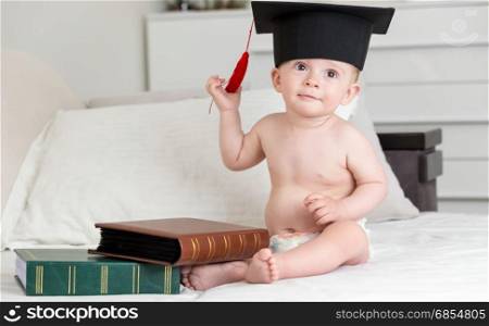 Baby boy siting on bed with books and taking off graduation cap
