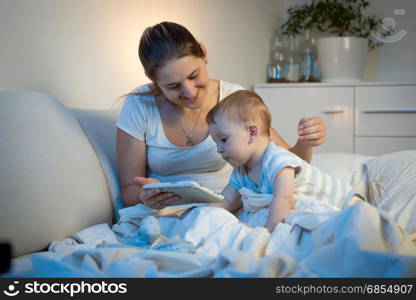 Baby boy sitting on bed with mother and using digital tablet at night