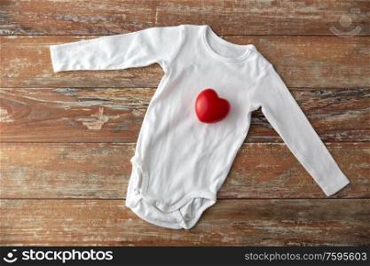 baby clothes, babyhood and clothing concept - bodysuit with red heart toy on wooden table. baby bodysuit with red heart toy on wooden table