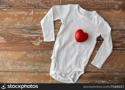 baby clothes, babyhood and clothing concept - bodysuit with red heart toy on wooden table. baby bodysuit with red heart toy on wooden table