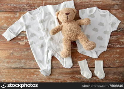 baby clothes, babyhood and clothing concept - bodysuits and socks with teddy bear toy on wooden table. baby bodysuits and teddy bear on wooden table