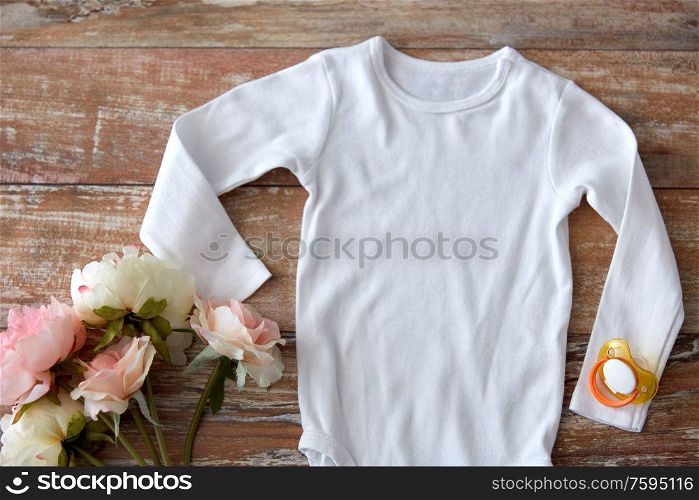 baby clothes, babyhood and clothing concept - white bodysuit with soother and flowers on wooden table. baby bodysuit with soother and flowers on wood