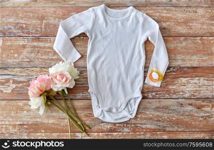 baby clothes, babyhood and clothing concept - white bodysuit with soother and flowers on wooden table. baby bodysuit with soother and flowers on wood