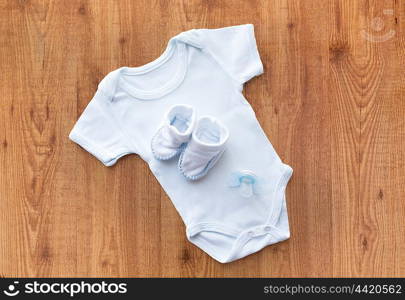 baby clothes, babyhood, motherhood and object concept - close up of white bodysuit, bootees and soother for newborn boy on wooden table