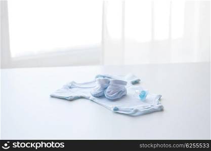 baby clothes, babyhood, motherhood and object concept - close up of white bodysuit, bootees and soother for newborn boy on table at home or maternity hospital