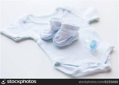 baby clothes, babyhood, motherhood and object concept - close up of white bodysuit, bootees and soother for newborn boy on table