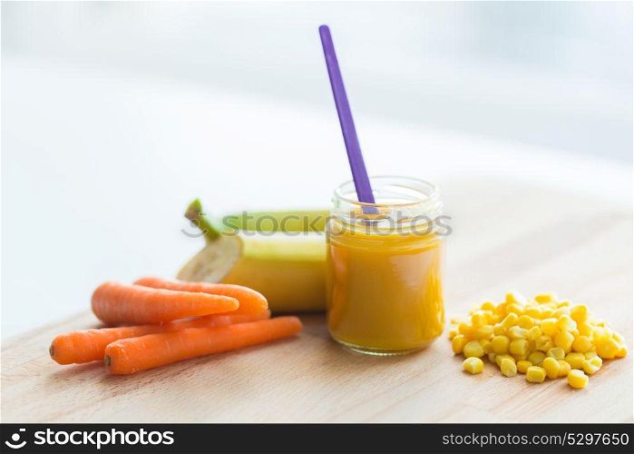 baby food, healthy eating and nutrition concept - glass jar puree with carrot, banana and corn on wooden board. puree or baby food with fruits and vegetables