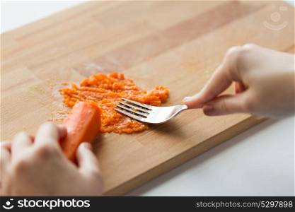 baby food, healthy eating and nutrition concept - hand with fork making mashed carrot on wooden board. hand with fork making mashed carrot on board