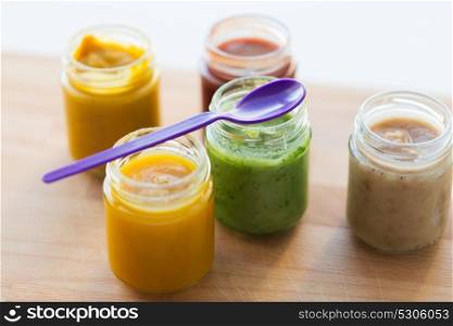 baby food, healthy eating and nutrition concept - open glass jars of vegetable or fruit puree and feeding spoon on wooden board. vegetable or fruit puree or baby food in jars