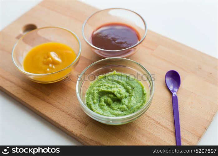 baby food, healthy eating and nutrition concept - vegetable or fruit puree in glass bowls and feeding spoon on wooden board. vegetable puree or baby food in glass bowls
