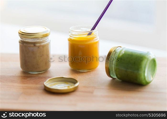 baby food, healthy eating and nutrition concept - vegetable or fruit puree in glass jars and feeding spoon on wooden board. vegetable or fruit puree or baby food in jars