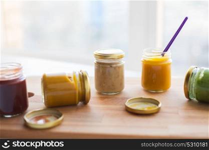 baby food, healthy eating and nutrition concept - vegetable or fruit puree or baby food in glass jars and feeding spoon on wooden board. vegetable or fruit puree or baby food in jars