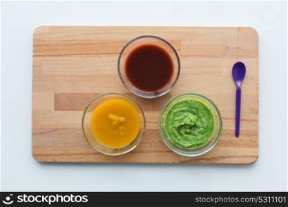 baby food, healthy eating and nutrition concept - vegetable or fruit puree in glass bowls and feeding spoon on wooden board. vegetable puree or baby food in glass bowls