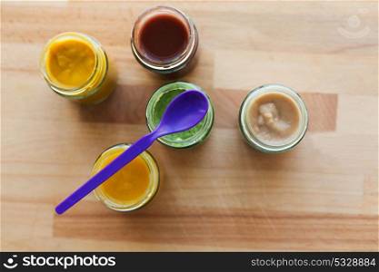 baby food, healthy eating and nutrition concept - vegetable or fruit puree or baby food in glass jars and feeding spoon on wooden board. vegetable or fruit puree or baby food in jars