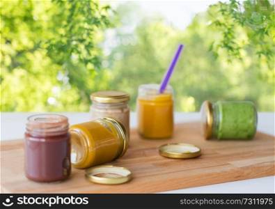 baby food, healthy eating and nutrition concept - vegetable or fruit puree in glass jars and feeding spoon on wooden board over green natural background. vegetable or fruit puree or baby food in jars
