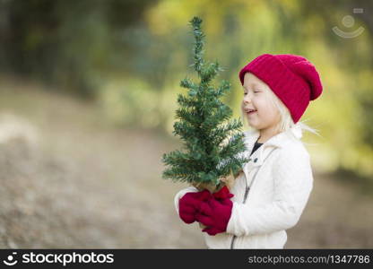 Baby Girl In Red Mittens and Cap Holding Small Christmas Tree Outdoors.