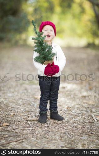 Baby Girl In Red Mittens and Cap Holding Small Christmas Tree Outdoors.