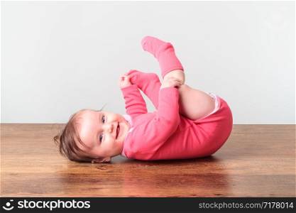 Baby girl lying on the floor and playing with her feet. She is looking at the camera. She is wearing a pink body suit. Space for text above