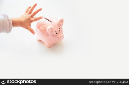 Baby hand reaching for savings in pink piggy bank. Bright white isolated background. Copy space, space for text.. Baby hand reaching for savings in pink piggy bank