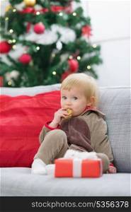 Baby in suit of Santa&rsquo;s little helper with Christmas gift eating cookie