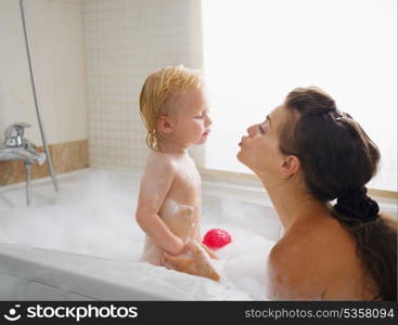 Baby kissing mother while washing in foam filled bathtub