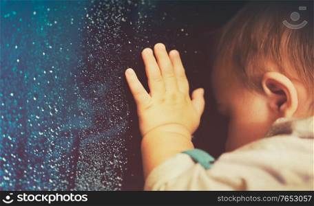 Baby looking through rainy window at home, vintage style photo of a little kid waiting for a parents from the work at the dark autumn evening