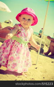 Baby with hat in hands of adult on sunny day. Child holding adult hands on beach