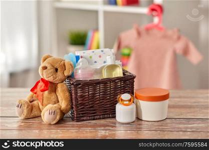 babyhood and care products concept - baby things in wicker basket and teddy bear toy on wooden table at home. baby things in basket and teddy bear toy on table