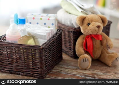 babyhood and care products concept - baby things in wicker baskets and teddy bear toy on wooden table at home. baby things in baskets and teddy bear toy on table