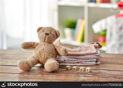 babyhood and clothing concept - baby clothes, teddy bear and toy blocks on wooden table at home. baby clothes and teddy bear toy on table at home
