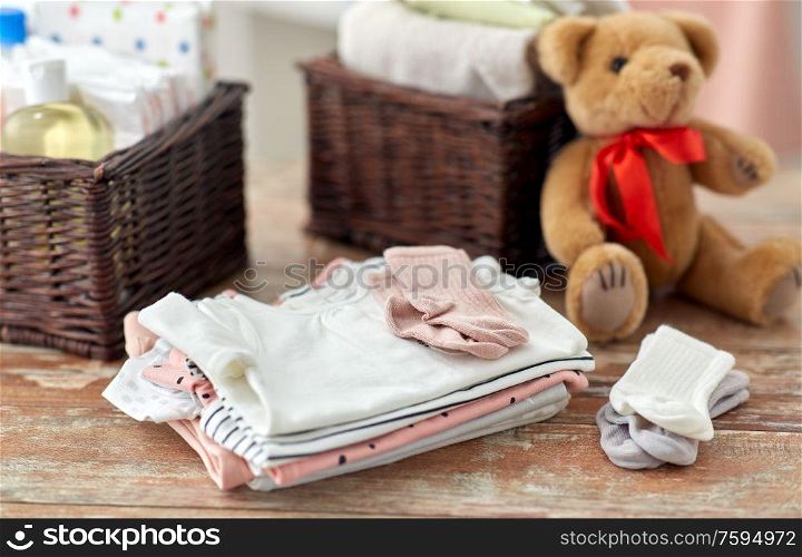 babyhood and clothing concept - baby clothes, teddy bear toy and baskets on wooden table at home. baby clothes and teddy bear toy on table at home